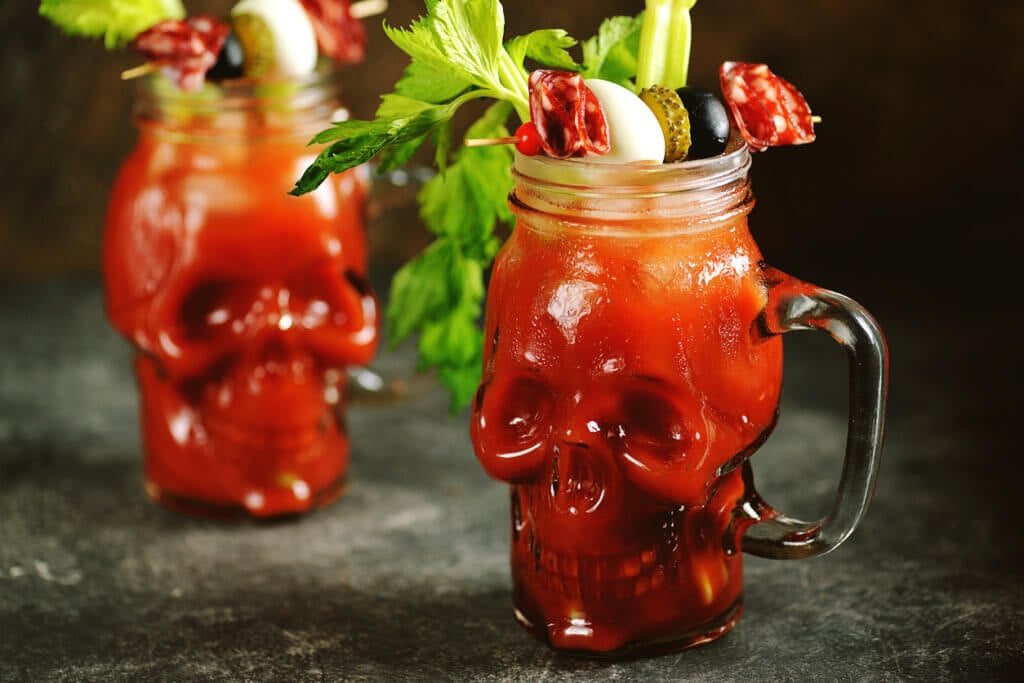 Bloody Mary cocktail in glass skull with celery sticks, pink salt, lime and canapes from canned vegetables. Halloween drink.