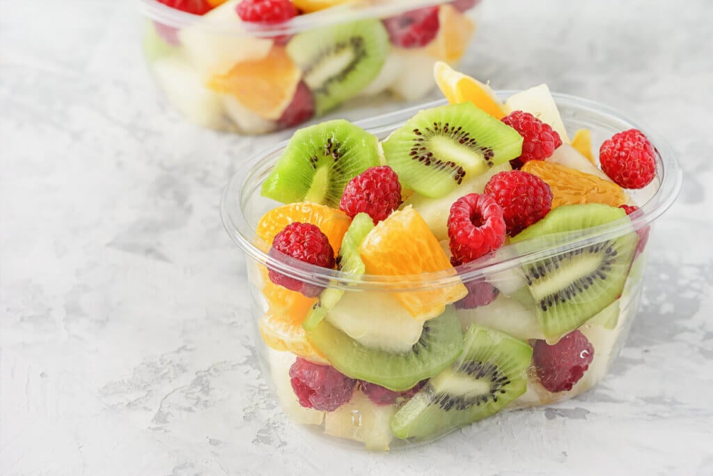 Fresh Sliced Tropical Fruits Berries in Container. Delicious Healthy Snack Ready to Eat Salad in Transparent Cup Closeup Elevated View. Vegetarian Raw Food. Citrus, Kiwi, Raspberries for Lunch