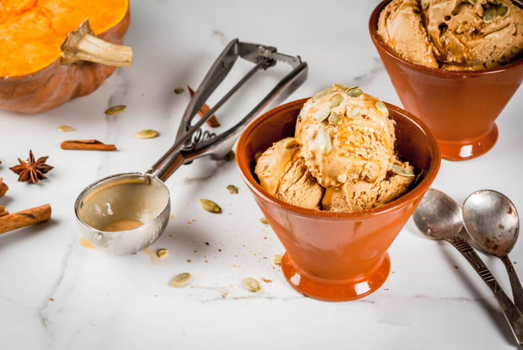 Ideas for autumn desserts, recipes from pumpkins. Pumpkin pie ice cream gelato in ceramic bowls, with maple syrup, pumpkin seeds, cinnamon and anise stars, on a white marble table.