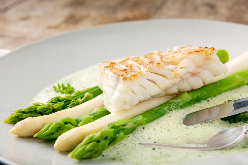 A dish of green and white asparagus topped with a piece of pan fried cod, surrounded by a parsley sabayonne foam.