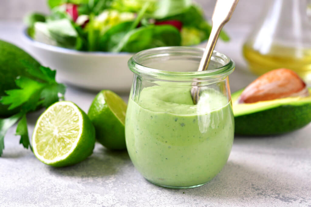 Homemade avocado yogurt dressing in a vintage glass jar with ingredients for making on a light slate, stone or concrete background.