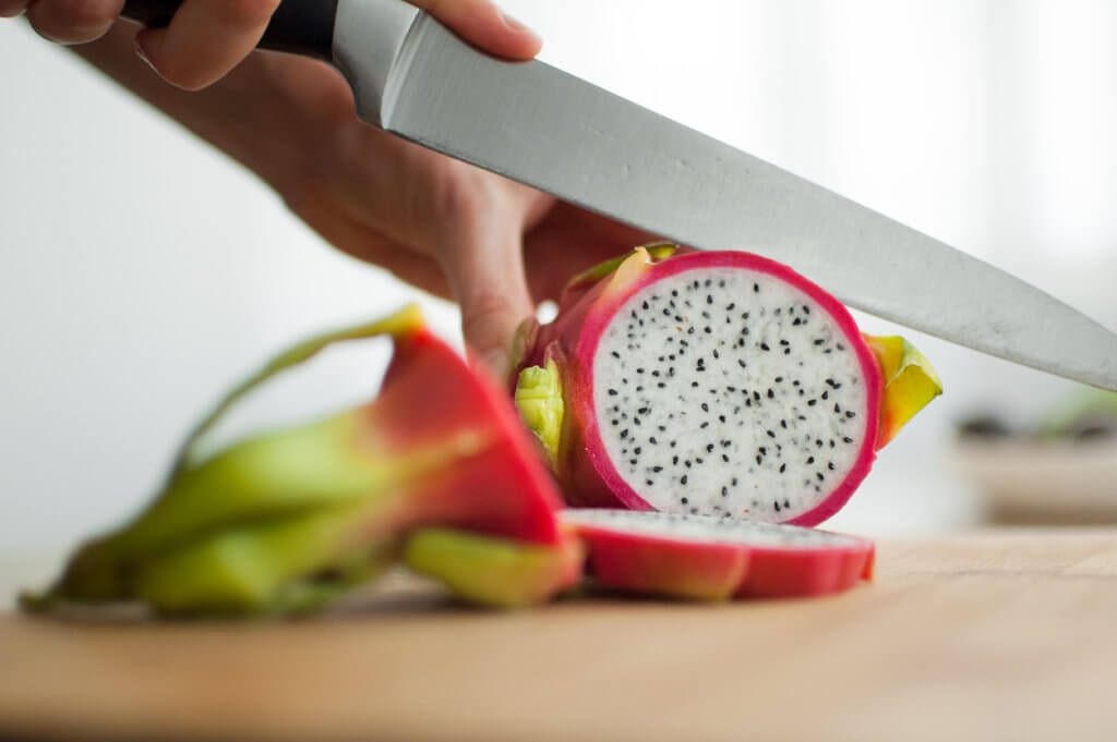 Hands cutting a dragon fruit or pitaya with pink skin and white pulp with black seeds on wooden cut board on the table. 