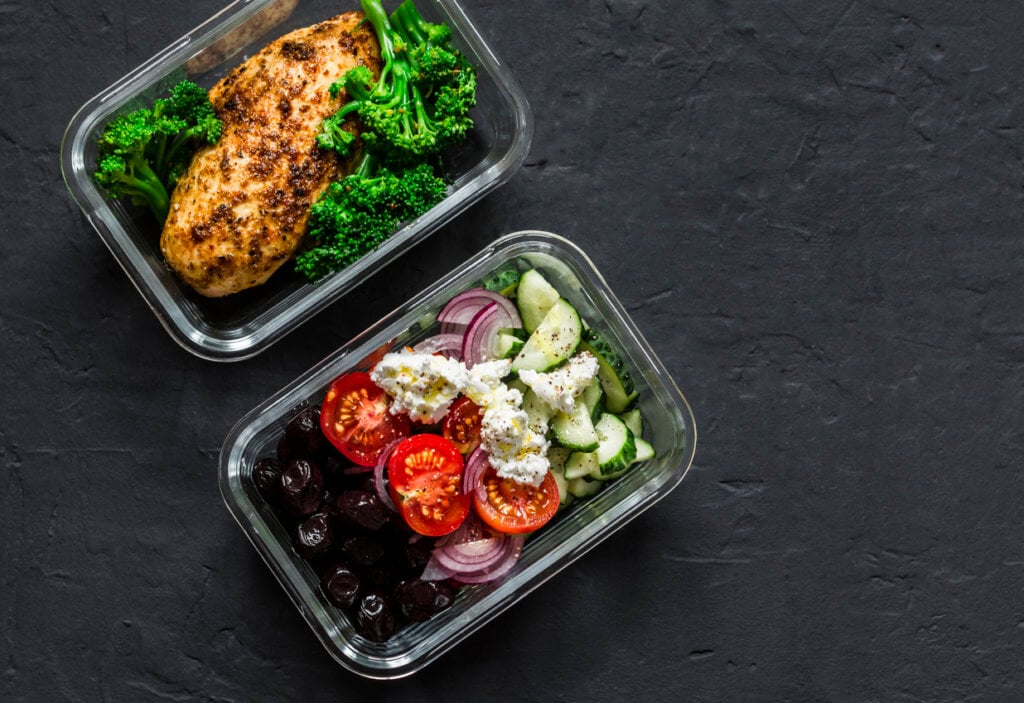Two healthy balanced lunch boxes with greek salad, baked chicken breast and broccoli on a dark background, top view