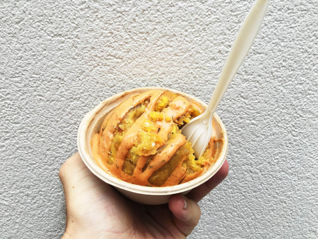 Hand holding a serving of Puerto Rican mofongo, fried plantains and garlic, with gourmet sauce in a sustainable paper street food bowl and a biodegradable fork.