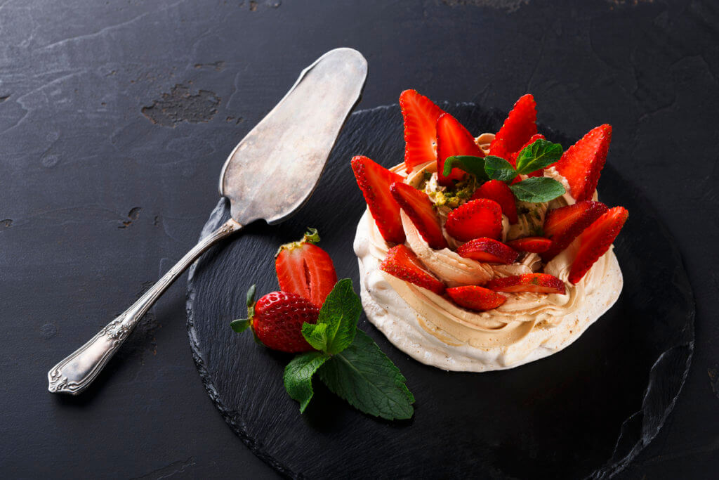 Refined dessert Pavlova made from meringue topped with low-fat whipped cream, mint leaves and strawberries on slate with cake spatula. 