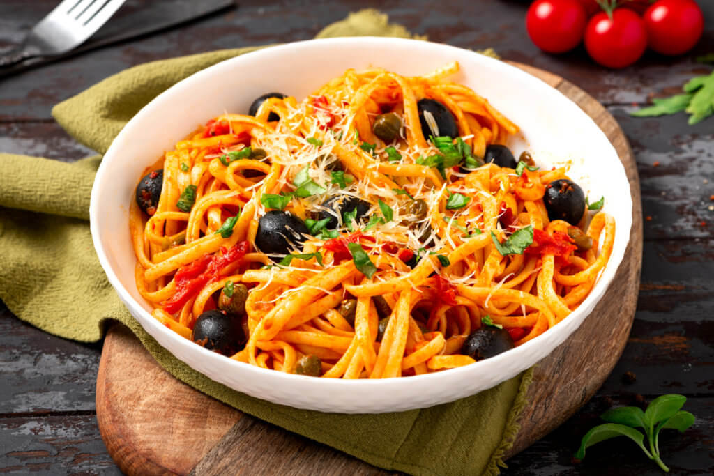 Pasta with anchovies, olives, capers, garlic, and tomatoes in a plate on a dark wooden background. 