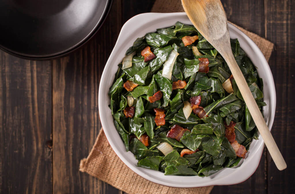 Cooked collard greens with bacon and onion on dark rustic farm table.