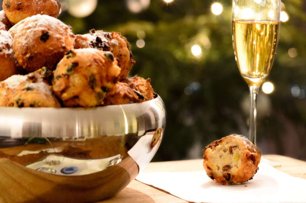 Oliebollen, oil balls or donut balls, a dutch pastry with raisins and powdered sugar traditionally eaten on New Year’s Eve in the Netherlands and a glass of champagne with a christmas tree in the background.