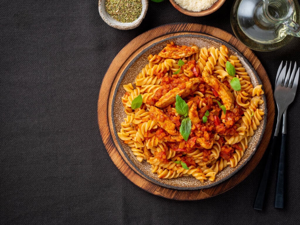 fusilli pasta with tomato sauce, chicken fillet with basil leaves on dark brown background.