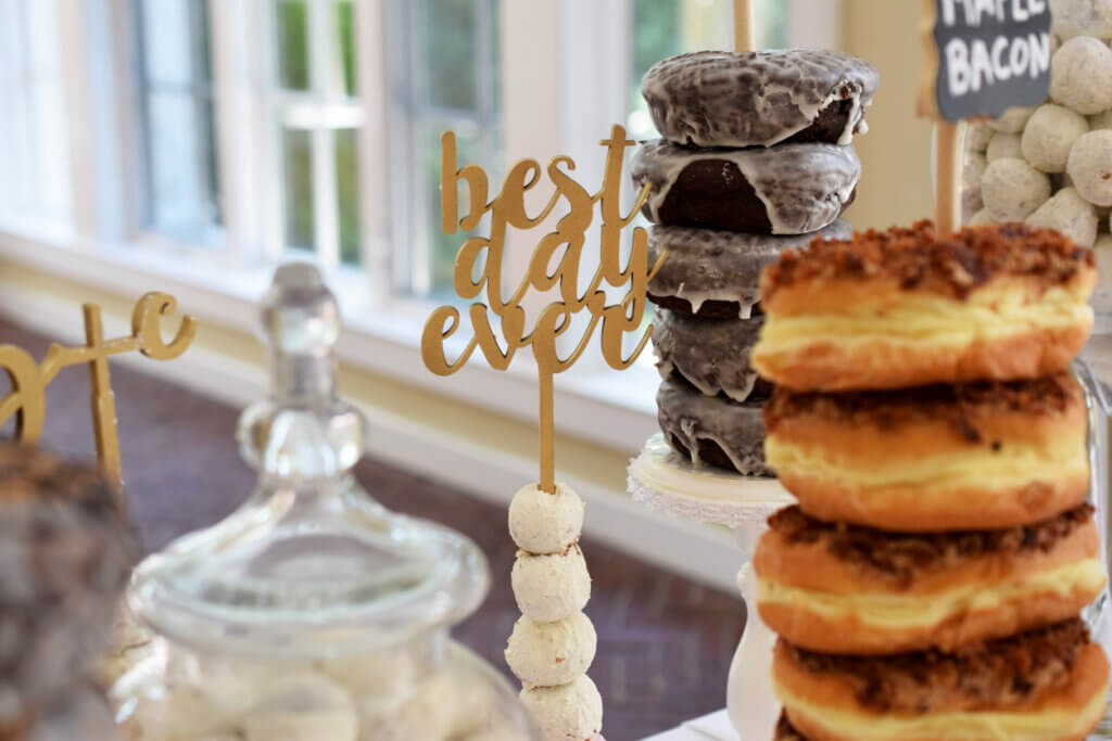 A gold “best day ever” sign at a wedding donut bar, surrounded by gourmet donuts in the foreground.