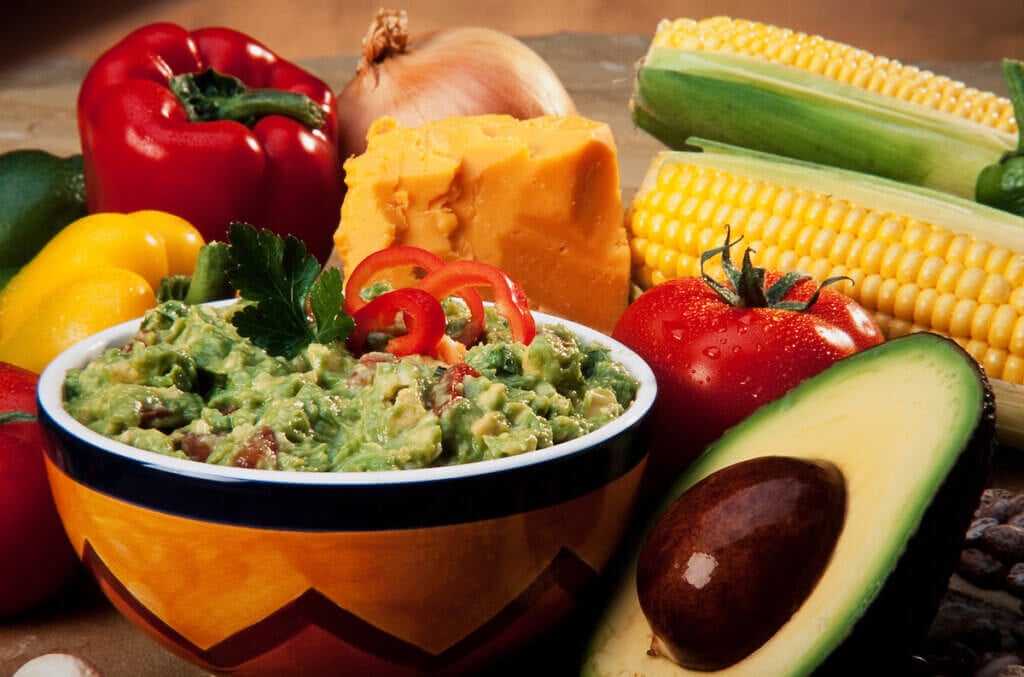 A bowl of guacamole surrounded by ingredients and other taco making ingredients