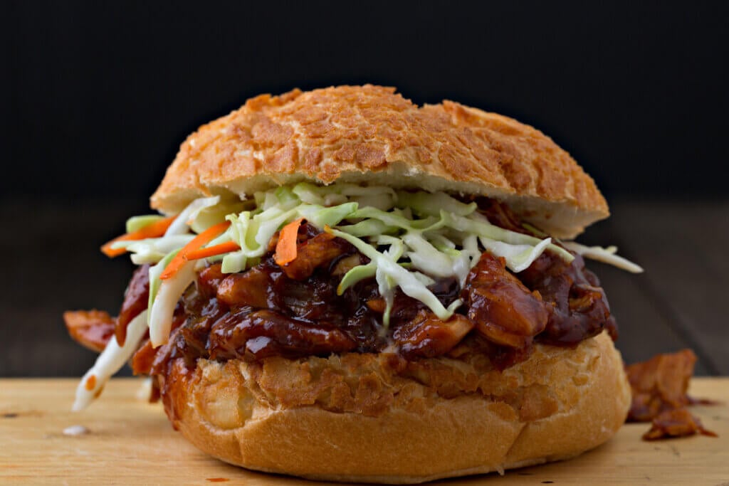 An extreme close up horizontal photograph of a barbecued, pulled chicken sandwich topped with freshly made crisp coleslaw in a dutch crust bun. 