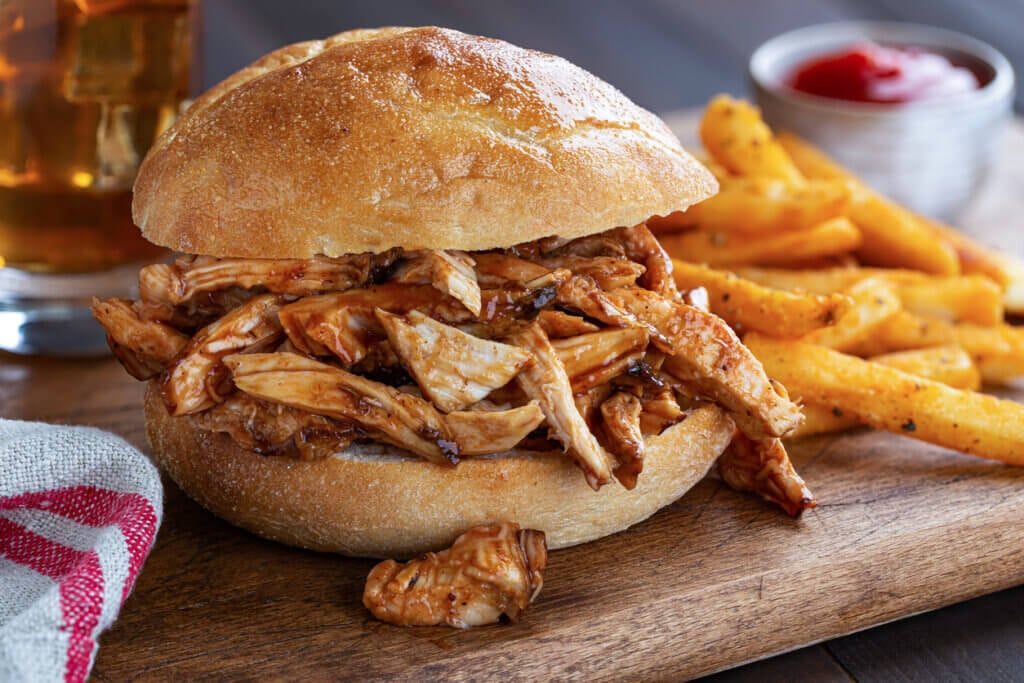 Closeup of a barbecue chicken sandwich on a bun with french fries in background on a rustic wooden cutting board