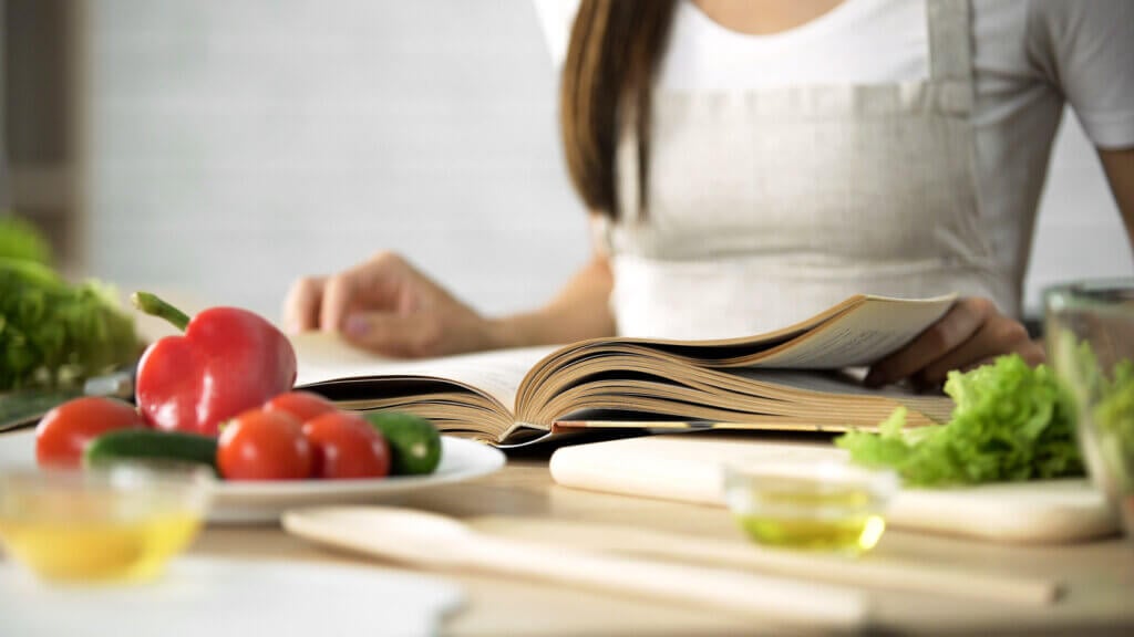 Reading cooking book with fresh vegetables and kitchen tools on table