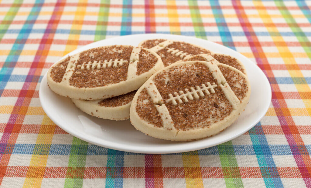 A plate full of football shaped sugar cookies with sprinkles on a colorful place mat.