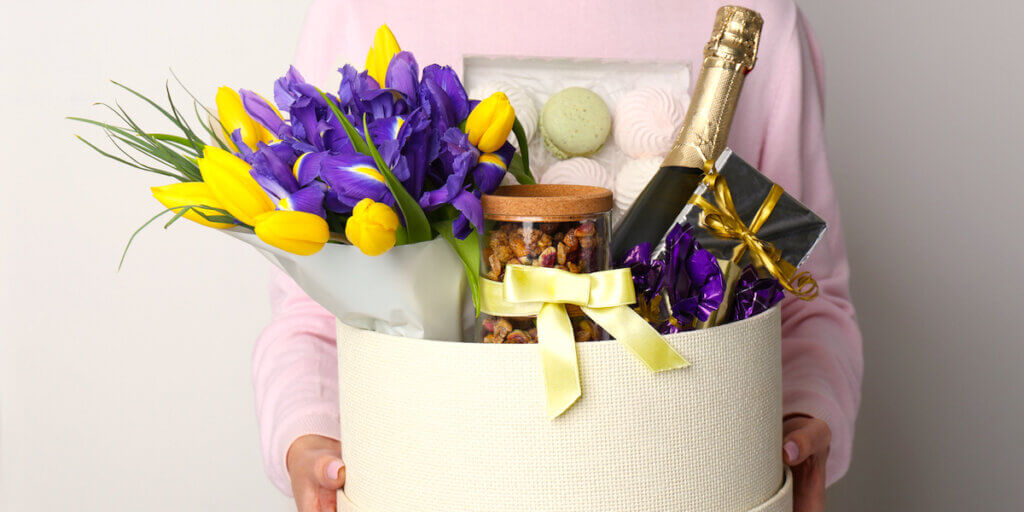 Woman holding basket with gifts, bouquet and champagne on light grey background