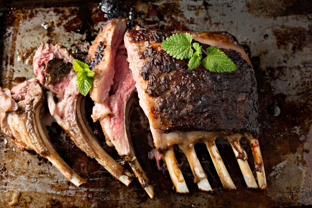 An overhead, close up horizontal, photograph of a baking tray with a cooked rack of lamb with several ribs sliced off.