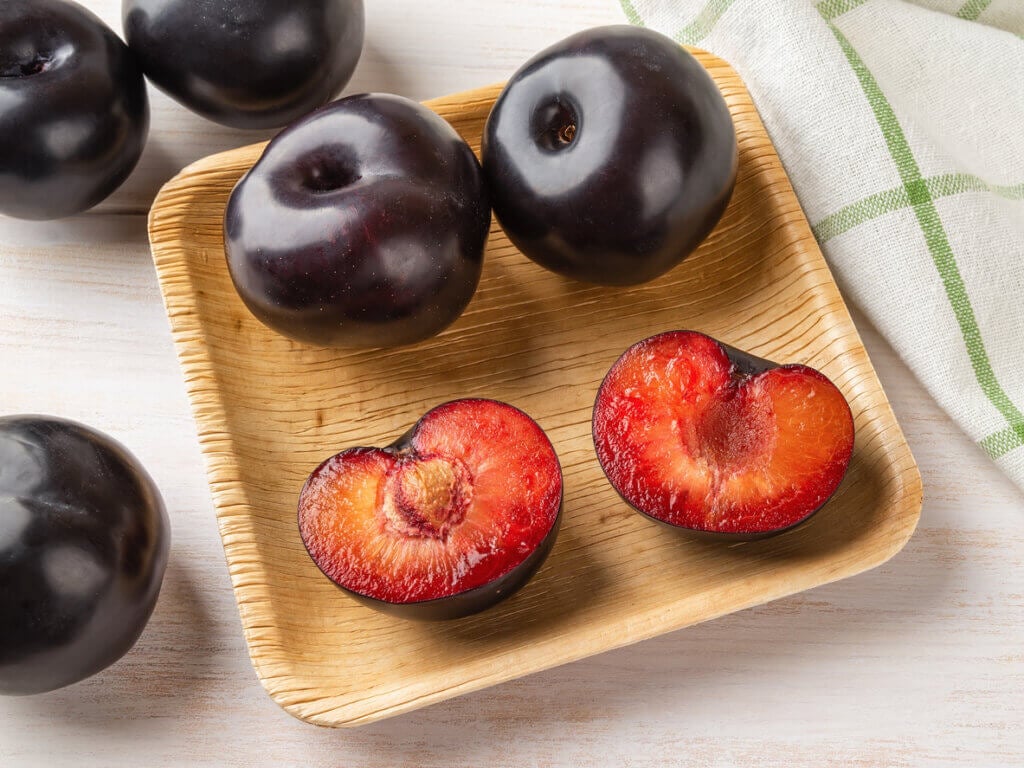 Black plums whole and cut in half on a square wooden plate.