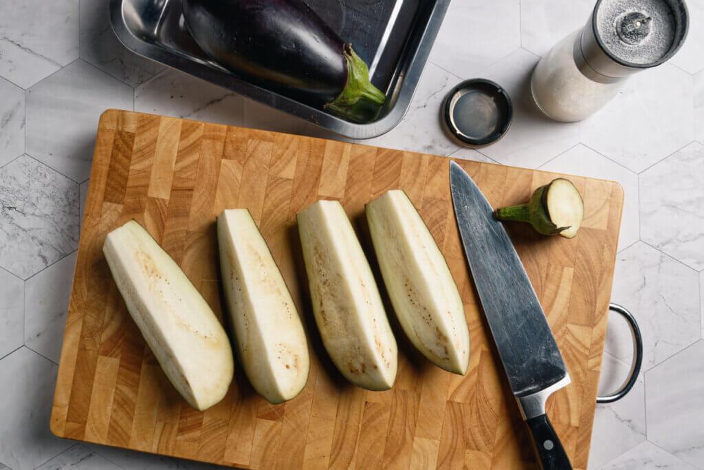 Eggplant cut into quarters lengthwise. With a chef's knife on a wooden cutting board.