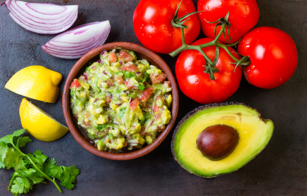 Guacamole and ingredients - avocado, tomatoes, onion, cilantro on a dark background.