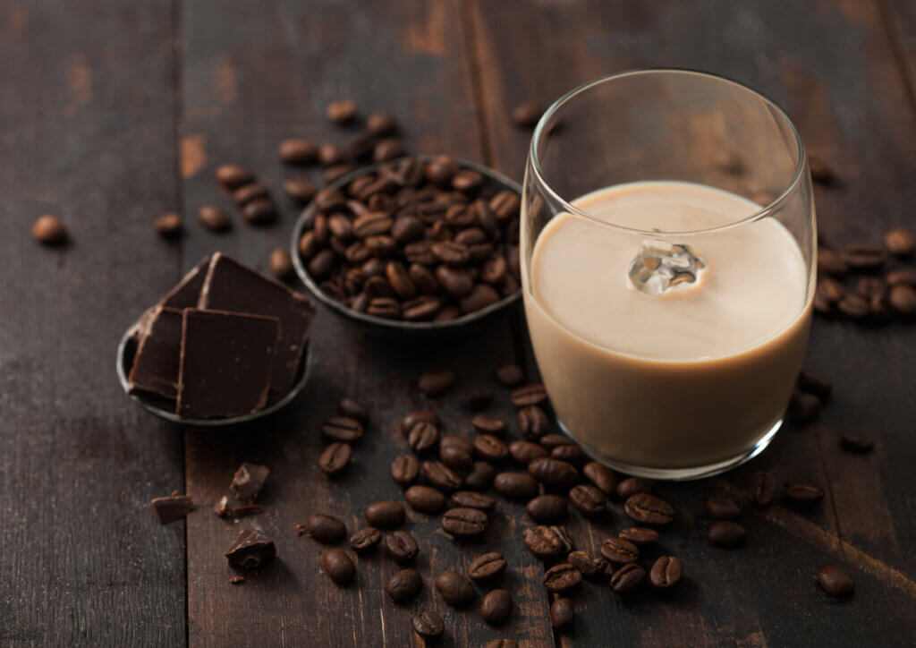 Glass of Irish cream liqueur with coffee beans and dark chocolate in steel bowl on dark wood background.
