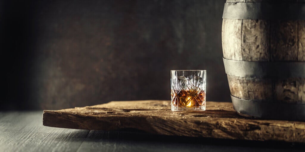Glass of bourbon in ornamental glass next to a vinatge wooden barrel on a rustic wood and dark background