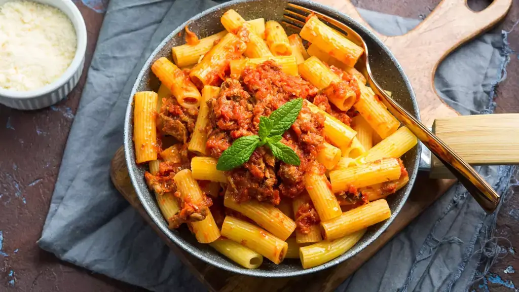 Bowl of rigatoni chickpea pasta with meat sauce on top next to a small ramekin with Parmesan cheese